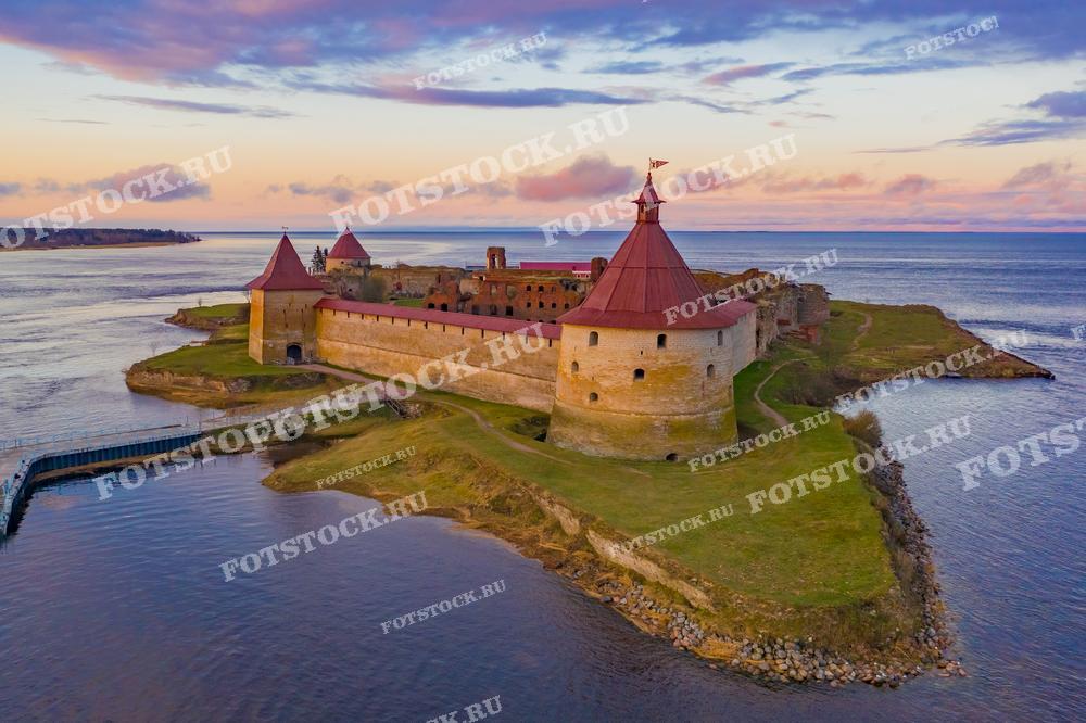 На фотографии запечатлен величественный вид на крепость Орешек, расположенную на острове в устье реки Невы. Закатное небо окрашивает облака в мягкие оттенки розового и голубого, создавая контраст с красными кирпичами и зелеными лужайками крепости. Это место является историческим памятником и популярной туристической достопримечательностью, фотография подойдет для использования в туристических брошюрах, образовательных материалах и публикациях, посвященных истории и архитектуре.